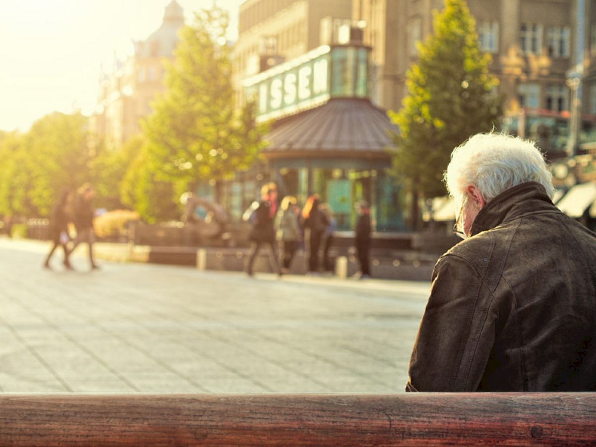 An elderly person sits on a bench in a sunlit urban area, with people walking in the background near a building labeled 