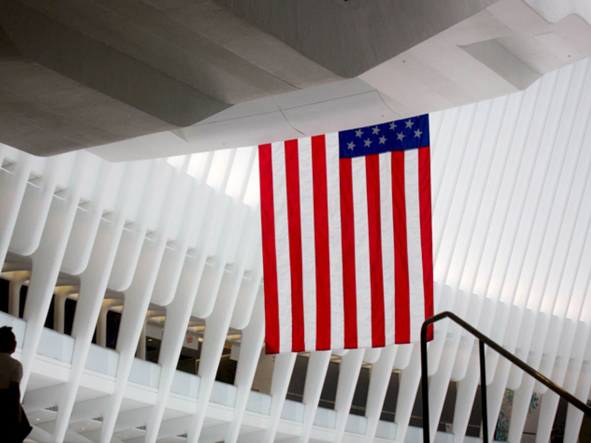 The image shows an interior with a large American flag hanging from the ceiling. A person is seen walking below, and the architecture features white vertical beams.